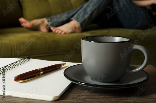 Gray cup of coffee  blank notebook and pen on the desk  woman relaxing on the green sofa.Concept of work and rest