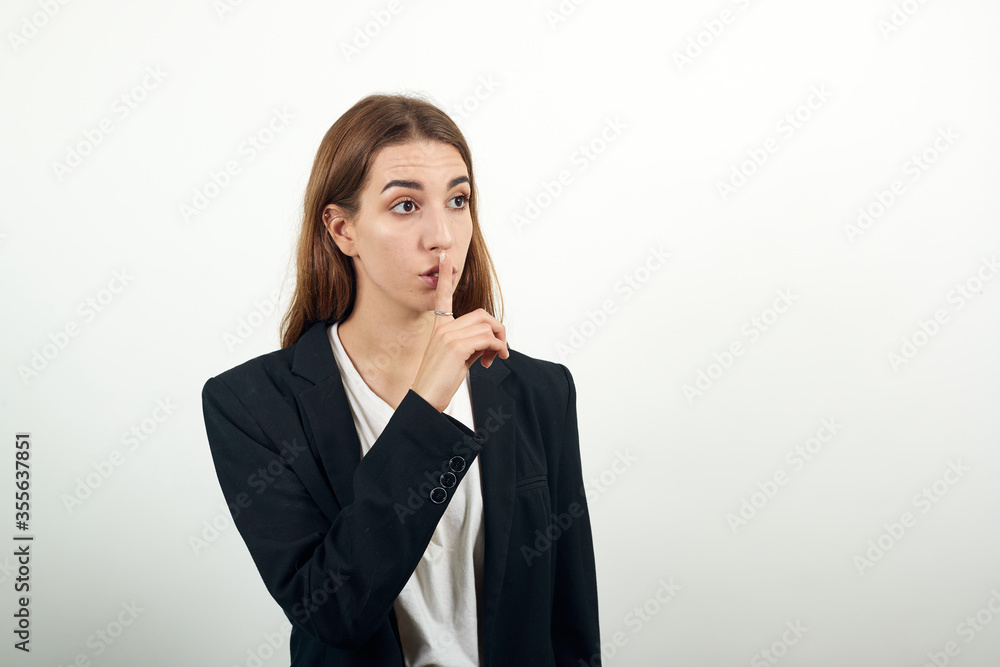 Index finger on lips, silence gesture, shhh quiet, asks for voicelessness forefinger at the mouth. Young attractive woman with brown hair in a light t-shirt and black jacket on white background Index finger on lips, silence gesture, shhh quiet, asks for voicelessness forefinger at the mouth. Young attractive woman with brown hair in a light t-shirt and black jacket on white background