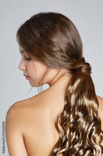 Cropped shot of a brown-haired lady with a wiglet made as a long chestnut ponytail with lustrous strands. The lady with naked shoulders is facing profile on the gray backdrop. 
