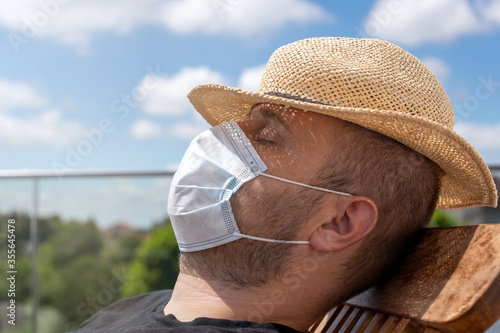 Sommerurlaub in Coronazeiten, Urlaub auf Balkonien mit Mundnasenschutz Maske - Mann mit Sonnenhut und Maske sonnt sich auf der Terrasse photo