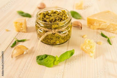 Single glass transparent jar with rope full of italian sauce basil pesto standing on the wooden table. Pieces of parmesan cheese, basil leaves and garlic lay on the light background. Close-up