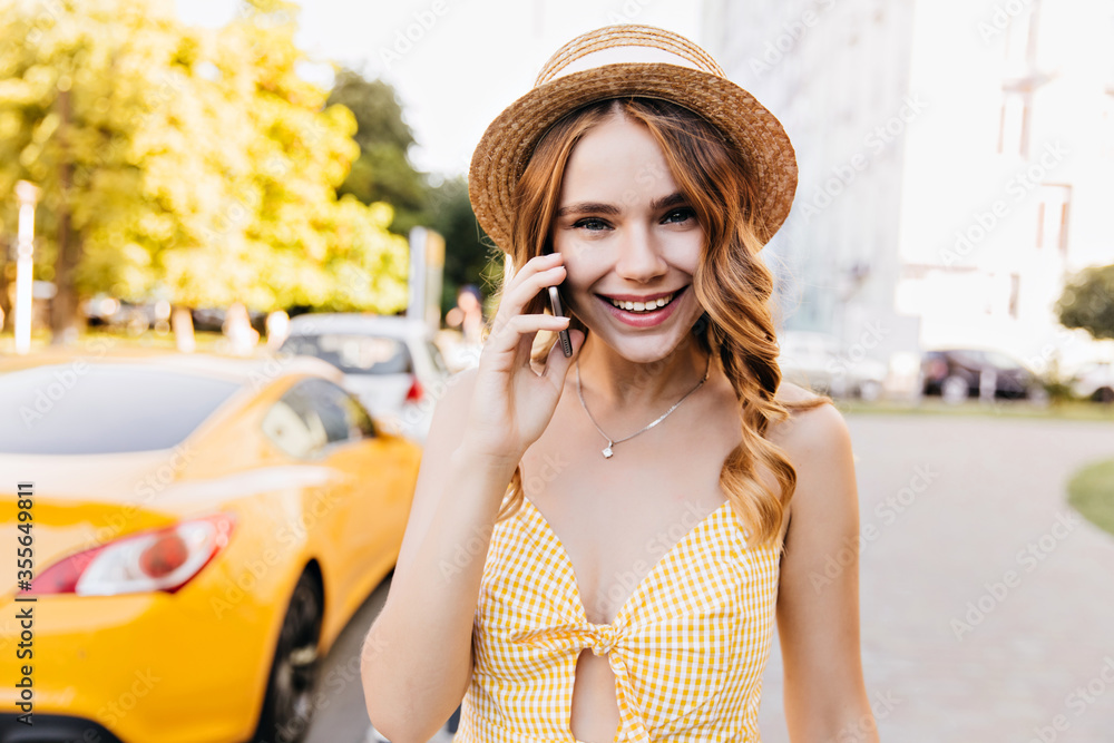 Elegant fair-haired girl in vintage hat talking on phone in summer weekend. Cute white female model in yellow attire enjoying morning walk.