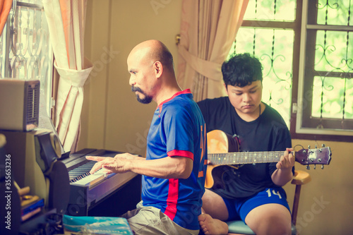 Selective focus to father playing piano with blurry Asian boy is playing guitar in home. Musical instrument for learning music, The music learning and enjoy concept.
