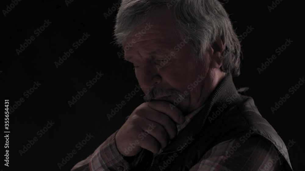 Concentrated grey haired senior man looking at the camera touching his chin standing in a dark studio isolated on a black background. Close up. Prores 422. Concentrated grey haired senior man looking at the camera touching his chin standing in a dark studio isolated on a black background. Close up. Prores 422.