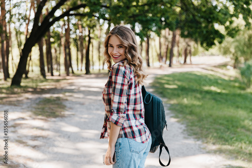 Glad girl in checkered shirt and blue jeans standing in park. Inspired woman with leather backpack smiling in spring day.