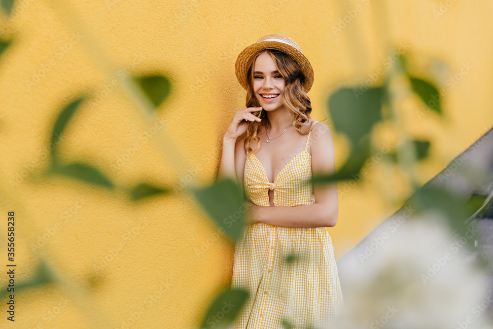 Outdoor portrait of lightly-tanned beautiful woman in hat isolated on yellow background. Graceful fair-haired girl in vintage dress smiling to camera.