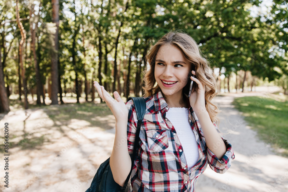 Pretty girl in good mood calling friend while walking in park. Outdoor shot of pleasant curly woman posing with backpack and phone.