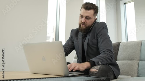 Young beard businessman work at the computer while is talking at phone about business. He is sitting on sofa in a business building. Work alone due the pandemic situation