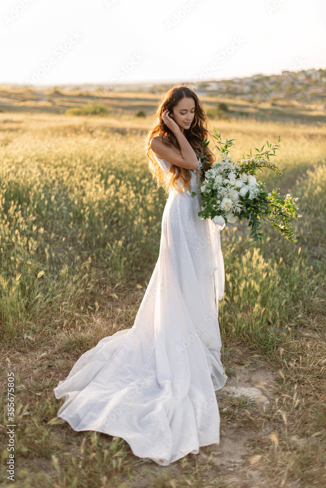 Refined slender bride in luxury fashion wedding dress holds a large wedding bouquet of peonies and wild flowers. Charming bride in a flowing dress stands in a green field of spikelets in the sunlight.