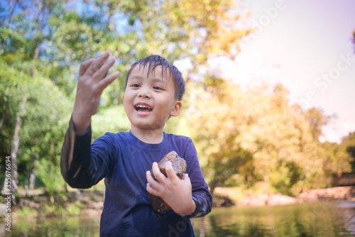 happy young Asia boy holding rocks in hands and smiling while standing in water pond at natural park during beautiful sunny day in summer season