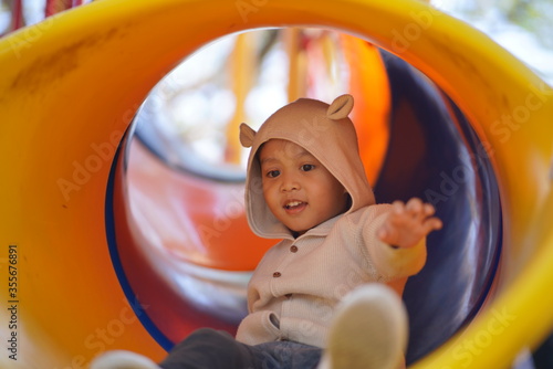 Cute small Asian boy playing in slider tunnel in public kid playground