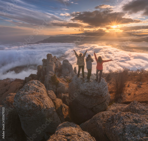 Tableau sur toile Girls tourists stand on a beautiful rock at a breakneck height above the clouds and watch amazing sunset in mountains