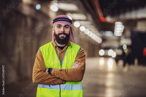 Cheerful smiling bearded arabian investor in vest, with headscarf on head standing in building in construction process with arms crossed.