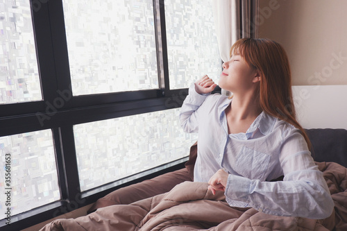 Image of a woman waking up and stretching in bed next to a bright window and in a cozy bed.
