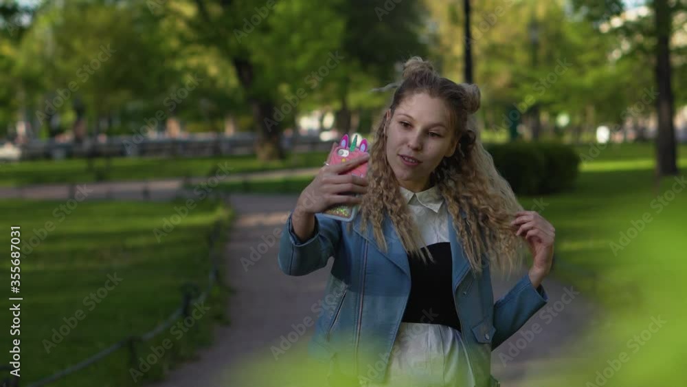Young beautiful girl with luxurious curly hair makes a video call by phone. Blonde is talking using the internet in a park. Young beautiful girl with luxurious curly hair makes a video call by phone. Blonde is talking using the internet in a park.