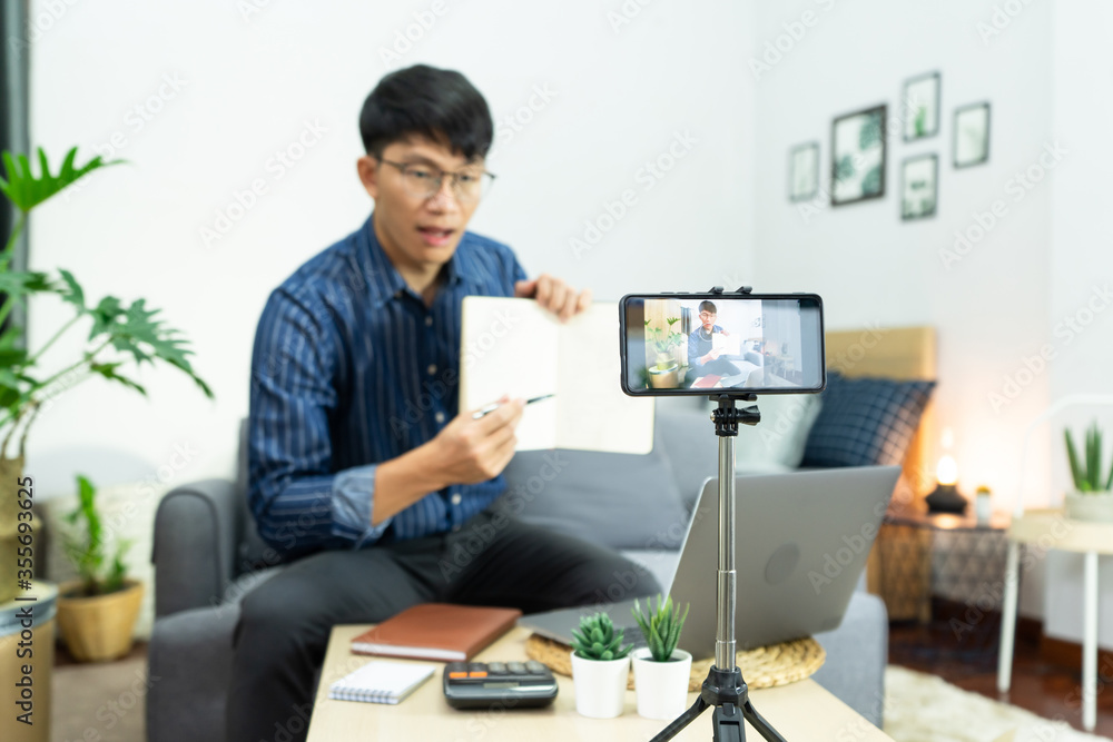 Young asian male blogger recording vlog video on camera review of product at home office, Focus on tripod mounted camera screen broadcast live stream video to a social network.