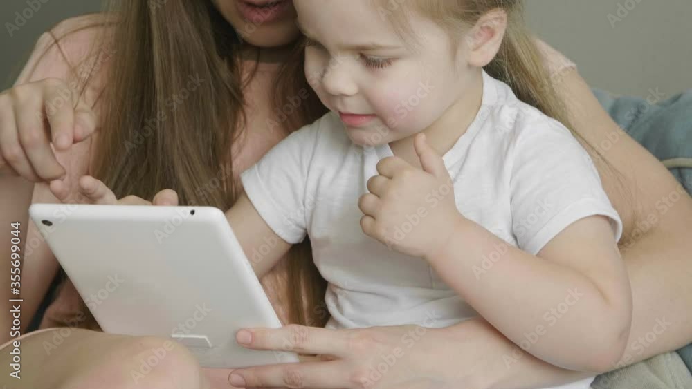 Mother with little cute daughter having fun and laughing using tablet. Funny games app. Mum with child take selfie, watch cartoons, make video call sitting on sofa at home. Mother with little cute daughter having fun and laughing using tablet. Funny games app. Mum with child take selfie, watch cartoons, make video call sitting on sofa at home.
