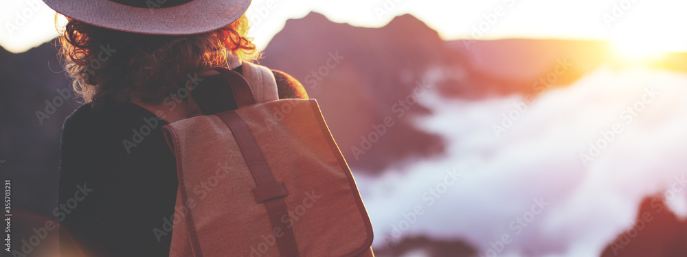 Young curly girl wearing  backpack and hat standing on edge of cliff and enjoying landscape, sunset, mountains and clouds. - obrazy, fototapety, plakaty 