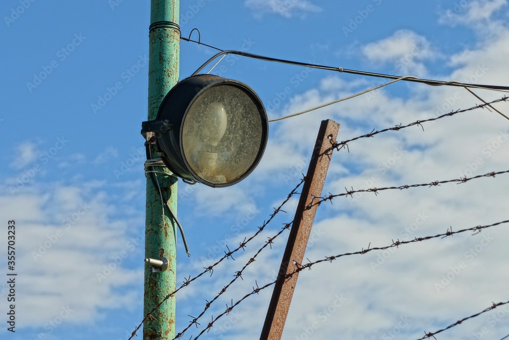 Obraz premium one old round spotlight with a lamp on an iron pole at the fence with barbed wire against the sky and clouds