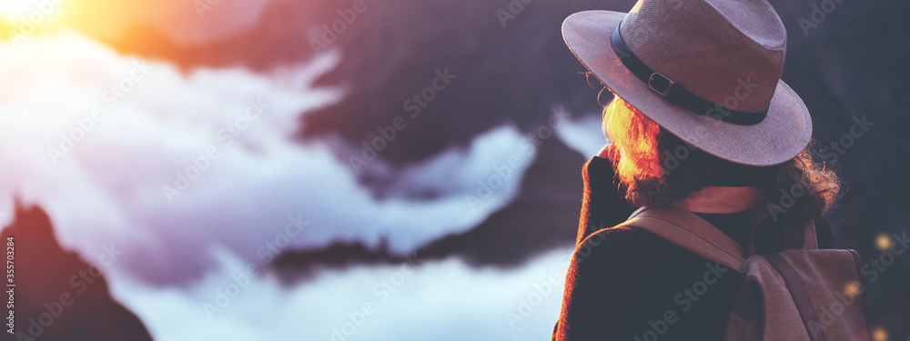 Young curly woman wearing  backpack and hat standing on edge of cliff and enjoying landscape, sunset, mountains and clouds. - obrazy, fototapety, plakaty 