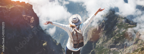 Young traveler woman with backpack and hat standing on edge of cliff in mountains at sunset against background of clouds.