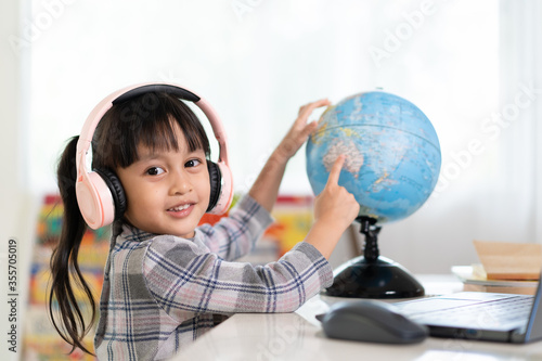 Asian beautiful little student girl is smiling and pointing on bilingual globe model for her online lesson, concept of learn from home and homeschooling for child education. photo