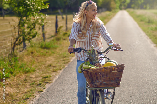 Happy woman smiling while riding a bicycle with a basket full of fresh and healthy fruits in a sunny day of summer in the countryside