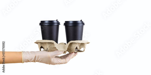 Two black disposable coffee cups on cardboard stand in gloved hand, isolated on white background. Concept of hygienic food preparation or home delivery. Banner, template, copy space