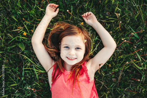 Cute smiling Caucasian girl resting in grass on meadow. Child lying on ground. Outdoors fun summer children activity. Kid having fun outside. Happy childhood lifestyle. View from top above.