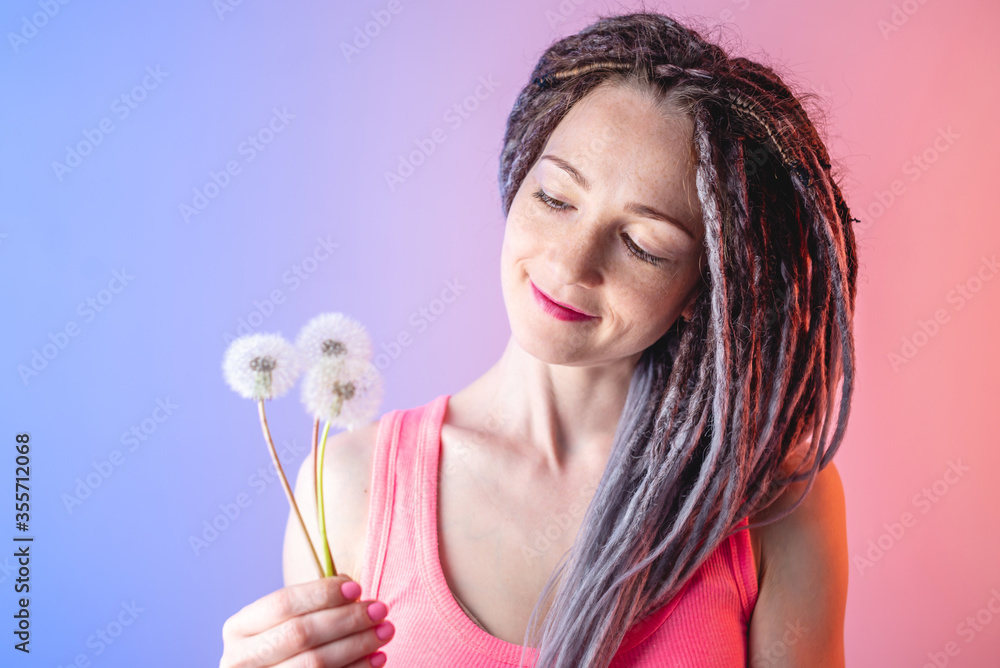 A young happy woman with dreadlocks is holding a white dandelion on a blue and pink background. Concept of a fun summer mood A young happy woman with dreadlocks is holding a white dandelion on a blue and pink background. Concept of a fun summer mood
