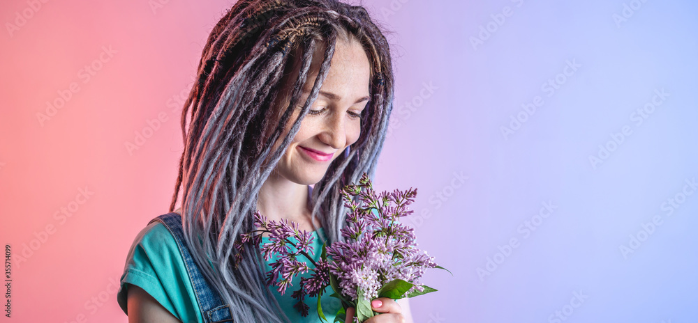 A young happy woman with dreadlocks is holding a beautiful bouquet of lilac on a blue and pink background. Concept of a bright cheerful mood