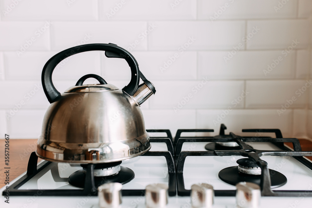 Metallic teapot on the gas oven. Bright kitchen interior. White modern dining room. Wooden complete kitchen with gas oven.