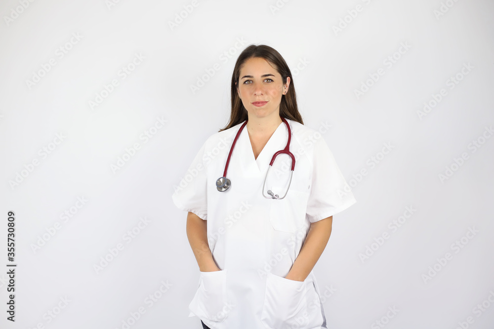 Young beautiful woman standing over white isolated background wearing her doctor uniform and smiling with her hands in her pocket Young beautiful woman standing over white isolated background wearing her doctor uniform and smiling with her hands in her pocket