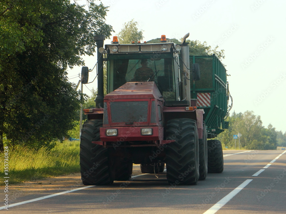 Big red heavy wheeled tractor with loaded green trailer drive on countryside asphalted road, rural transportation logistics, agricultural business, farming Big red heavy wheeled tractor with loaded green trailer drive on countryside asphalted road, rural transportation logistics, agricultural business, farming