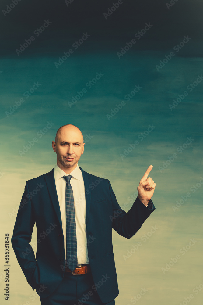 Young bald male businessman in a blue suit raised index finger up on a blue background. Looks to the camera. Young bald male businessman in a blue suit raised index finger up on a blue background. Looks to the camera.