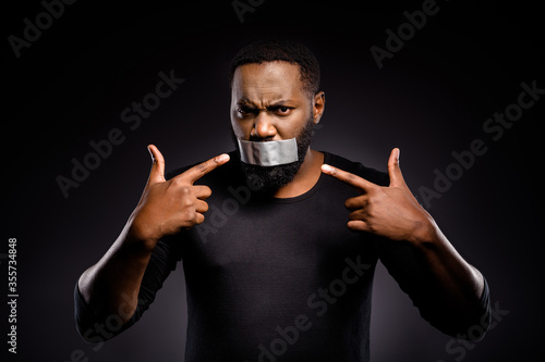 Photo of voiceless speechless afro american guy close cover adhesive tape lips point index finger suffer silence no talking racism injustice bully isolated black color background photo