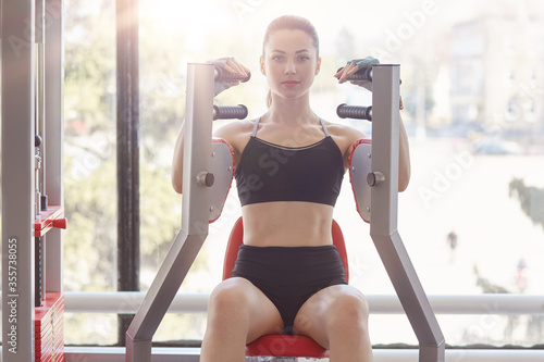 Young beautiful bodybuilder girl working out on fitness station in gym, training her biceps and triceps, having work out for upper body, wants develop definition of muscles. © sementsova321