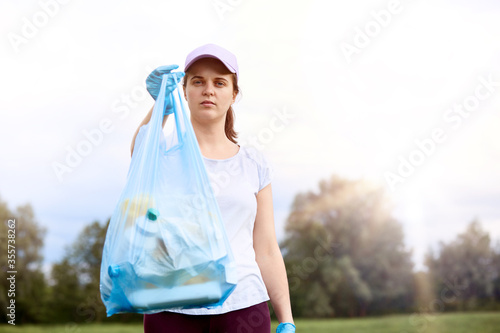 Young Caucasian girl wearing t shirt and baseball cap posing outdoor with garbage bag, cares for the environment, standing with sky and trees on background.