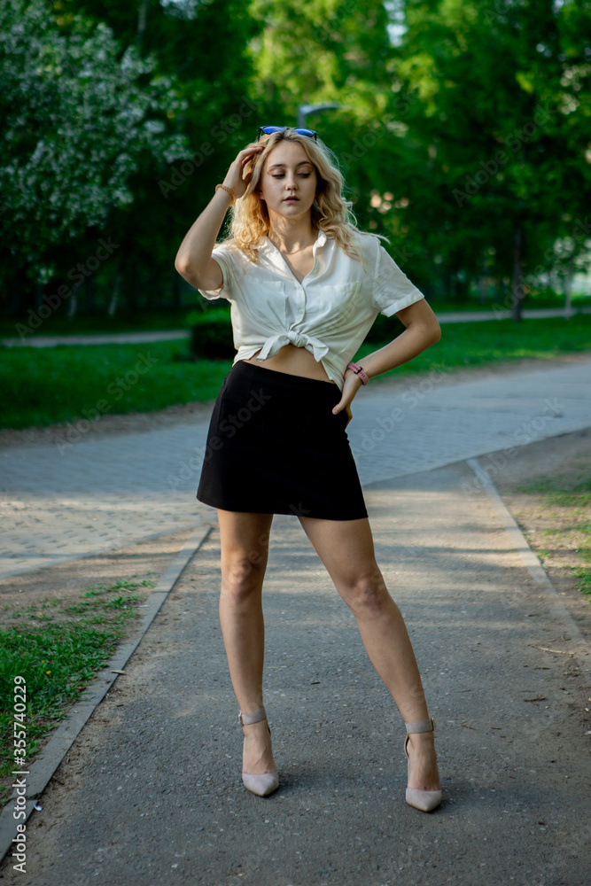 A teenage girl in a black skirt and white blouse. Light, elegant hair. Sunglasses. It stands in a Park on an asphalt path.