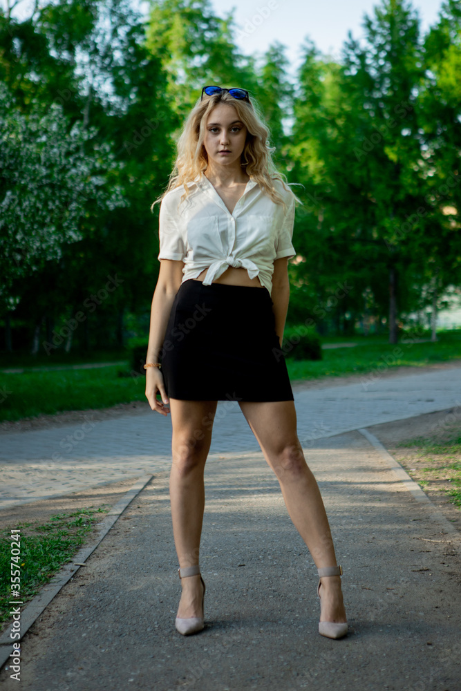 A teenage girl in a black skirt and white blouse. Light, elegant hair. Sunglasses. It stands in a Park on an asphalt path.