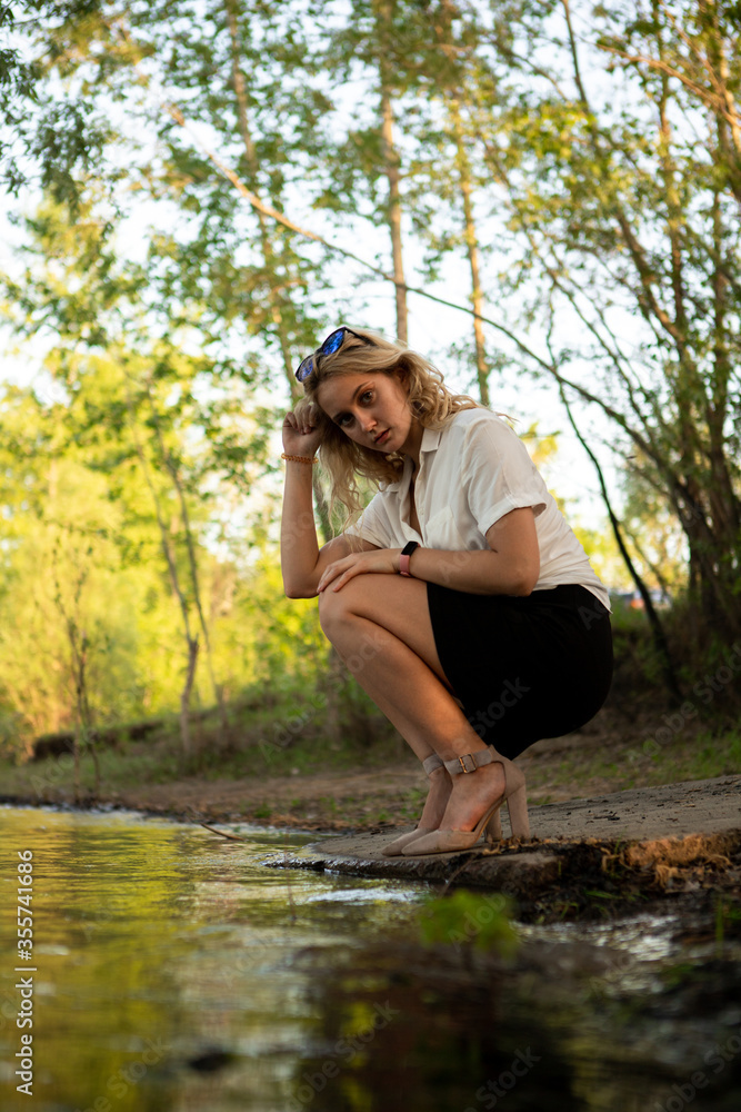 Girl near the water. A blonde woman in a white shirt poses on the Bank of a river or lake. Girl near the water. A blonde woman in a white shirt poses on the Bank of a river or lake.
