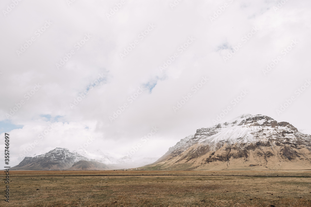 A field of dry yellow grass, against the backdrop of snow-capped mountain peaks in Iceland, in cloudy weather.