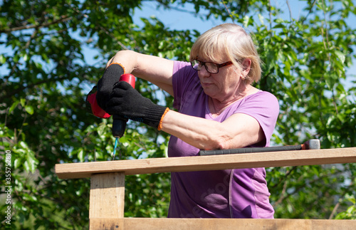 Senior Caucasian woman fastens a board on the roof of a farm building in the garden with a screwdriver