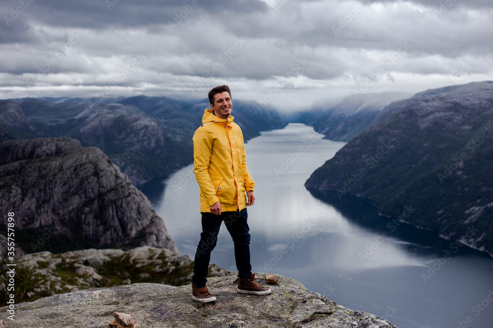 Brunet man hiker in yellow raincoat smiles and stays on the cliff of Preikestolen mountain (Preacher's Pulpit or Pulpit Rock) with background of Lysefjord and low cloudy sky in Norway
