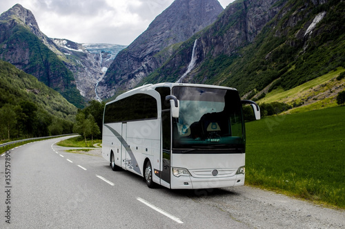 Trip to Norway. Blue ice tongue of Jostedal glacier melts from the giant rocky mountains into the green valley with a lot of waterfalls. Big white tourist bus rides on the road. 