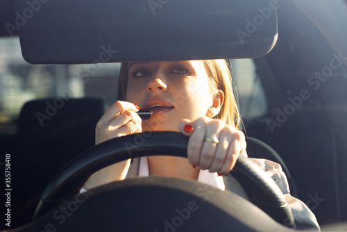 Woman driver sitting in car, doing make up using lipstick. Late for work in morning. Careless and dangerous driving. No concentration on road can cause fatal car accident. Stereotype for women driving photo