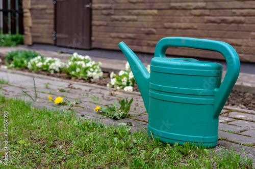 A green watering can for watering flowers and other plants stands next to a flower bed on a paving path.