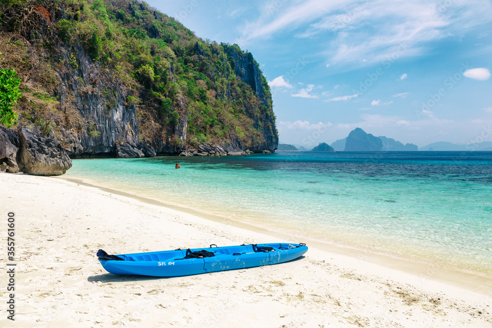 A blue kayak lies on white sand surrounded by mountains. Tropical beach with crystal clear water. A blue kayak lies on white sand surrounded by mountains. Tropical beach with crystal clear water.