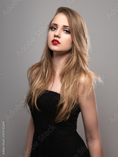 Beautiful bright makeup woman with long blond volume hair style in black dress, red lipstick looking on grey background.. Closeup
