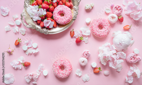 Top view composition of strawberry tray, three donuts with icing, marshmallows and pink flowers on a pastel pink background. Festive concept.